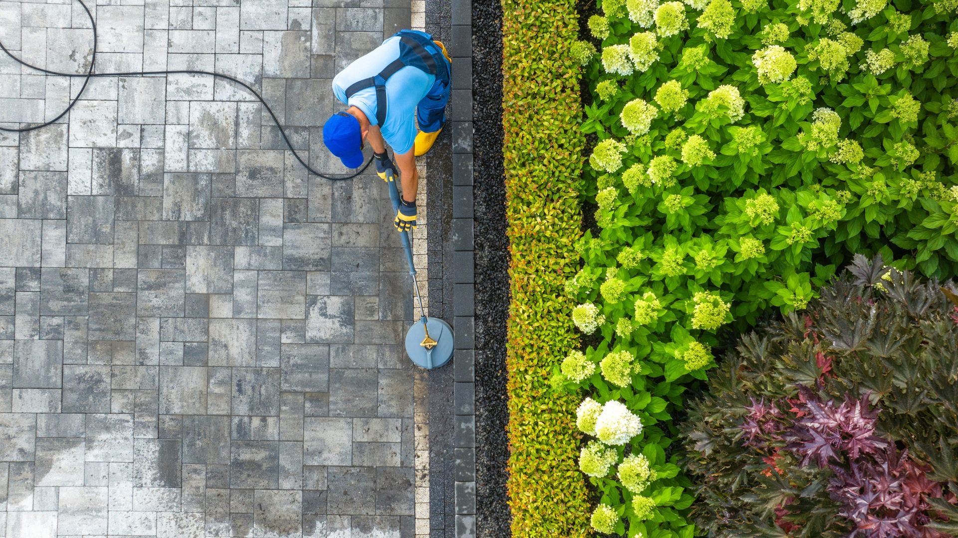 Worker pressure washing paved walkway next to lush garden landscaping